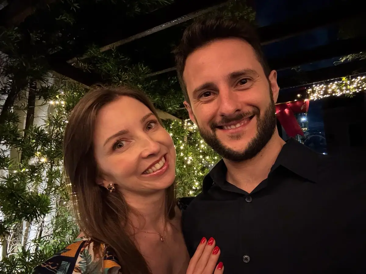 Cris e Kenni sorridente em ambiente externo com luzes decorativas ao fundo à noite