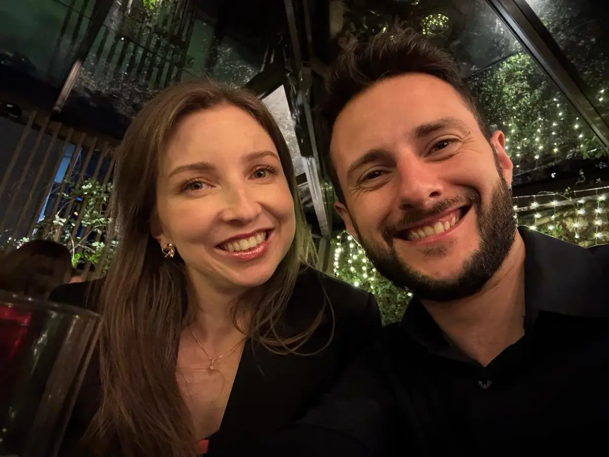 Cris e Kenni sorrindo em ambiente iluminado com luzes de fundo