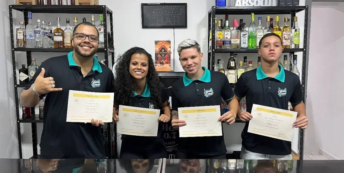 Quatro pessoas segurando certificados e sorrindo em frente a prateleiras com garrafas de bebidas em uma escola de bartenders.