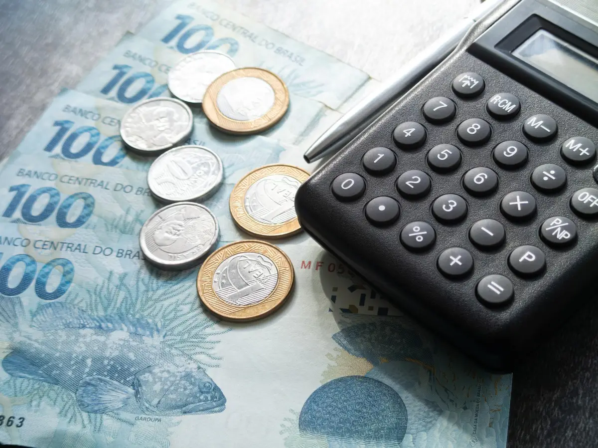 Notas de cem reais e moedas brasileiras ao lado de uma calculadora preta
