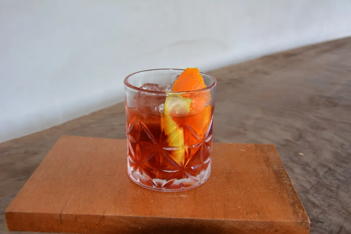 Copo com drink avermelhado, gelo e decoração de cascas de laranja e limão sobre tábua de madeira.