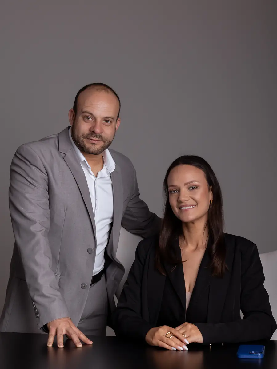 Homem de terno cinza em pé ao lado de mulher sentada vestindo paletó preto enquanto sorriem para a câmera