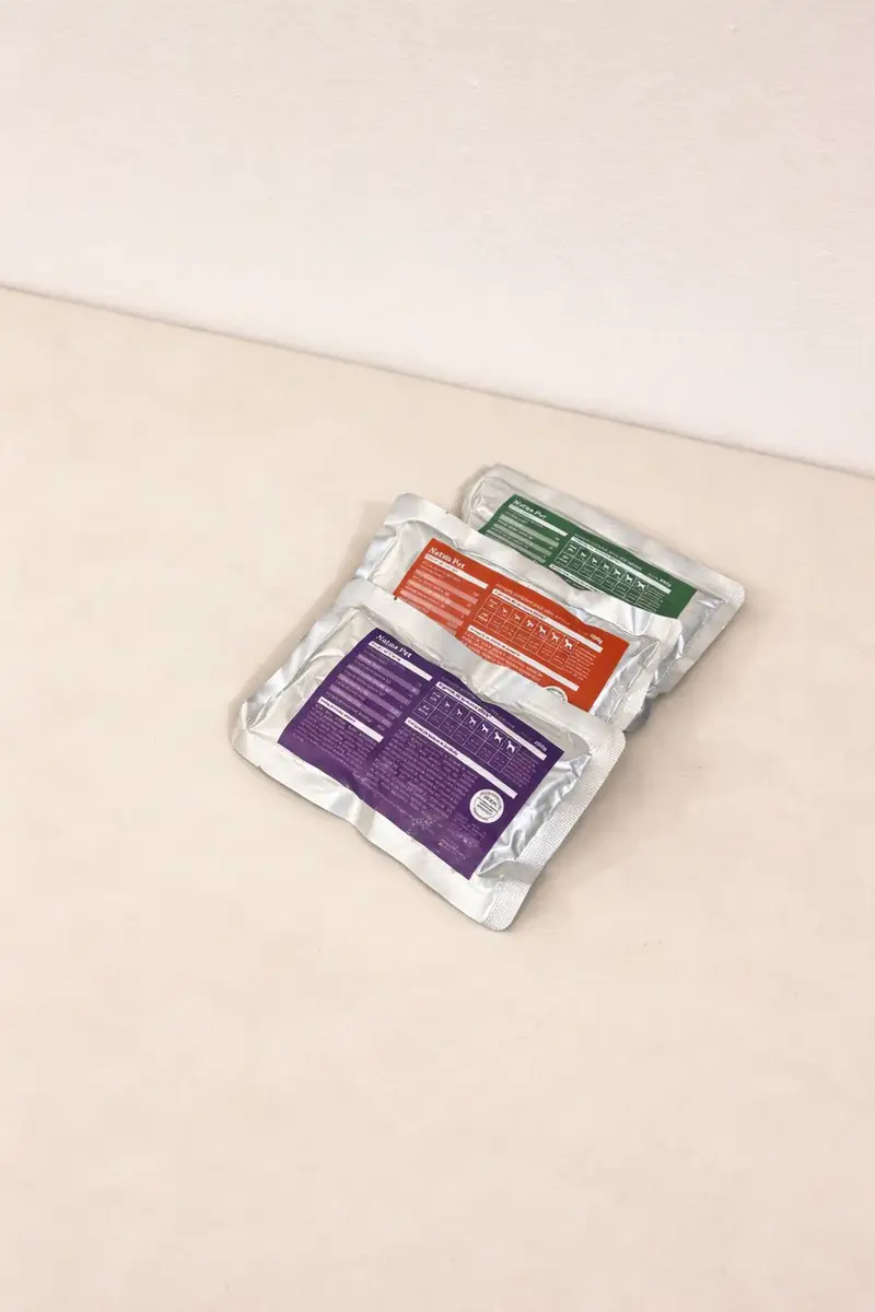 Três embalagens prateadas de comida natural para cães da Natua Pet com rótulos coloridos roxo, laranja e verde