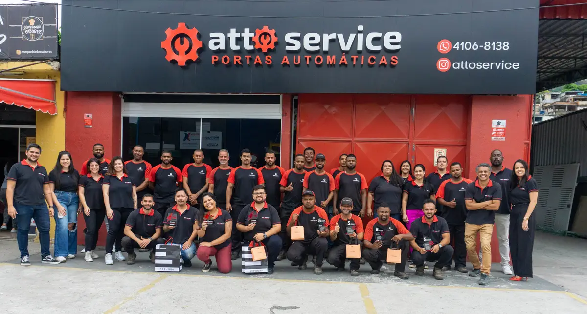 Equipe da Atto Service uniformizada em frente à sede da empresa com placas de contato ao fundo