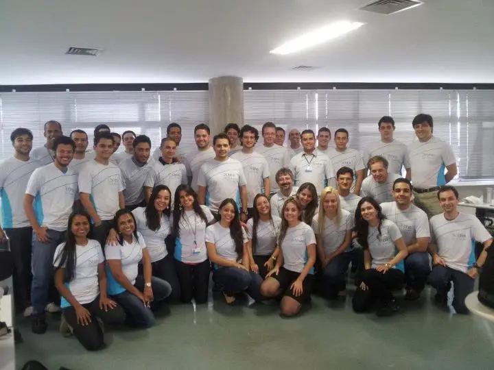 Grupo de cerca de 35 profissionais vestindo camisetas brancas posando em ambiente de escritório com janelas e persianas.
