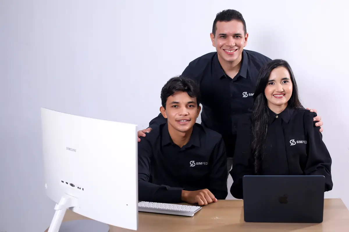 Três profissionais com camisas pretas da Simped sorrindo em mesa com computador e notebook
