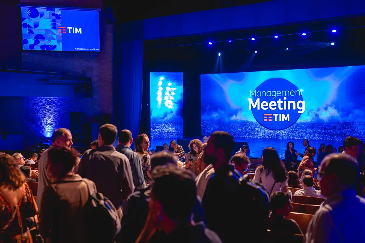 Pessoas reunidas em evento corporativo com palco iluminado em azul e tela grande com logo Management Meeting TIM