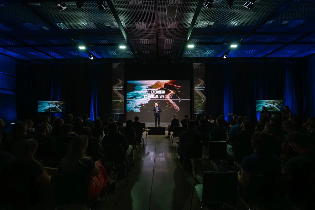 Palestrante em evento corporativo em auditório com público atento e tela grande ao fundo.