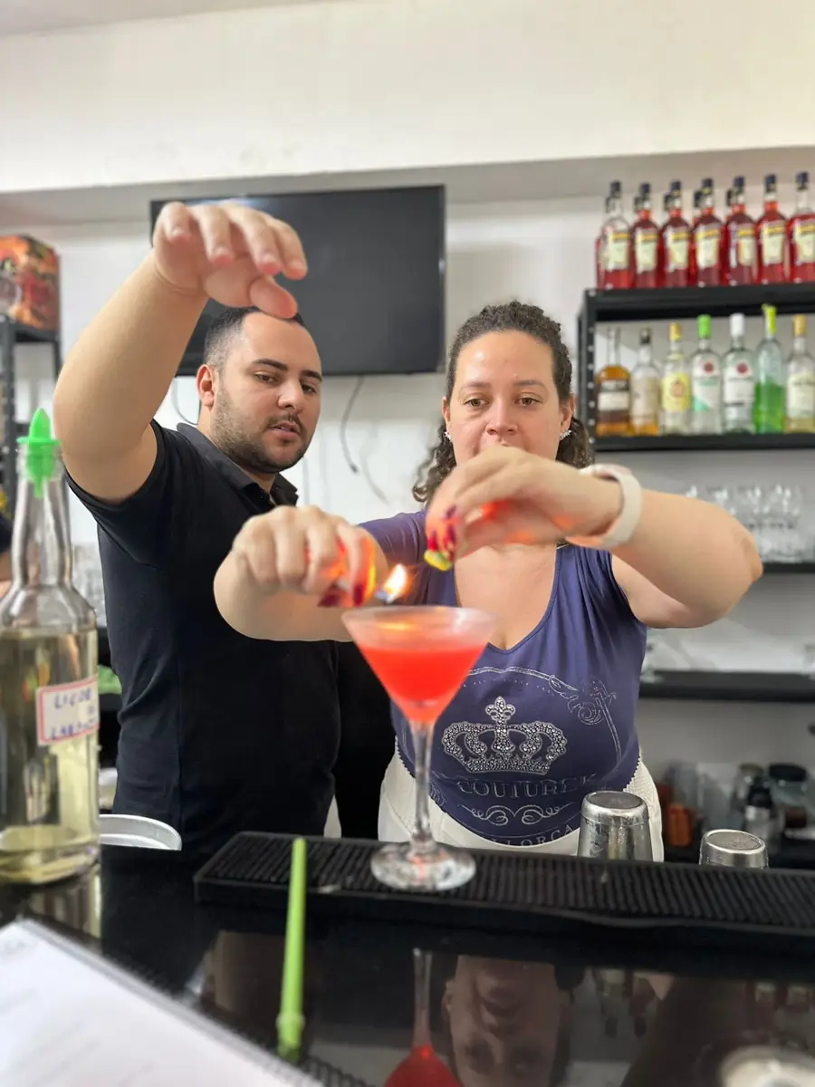 Pessoa acendendo a casca de um cítrico para flambear um drink vermelho em copo de cocktail, com outra pessoa orientando ao fundo
