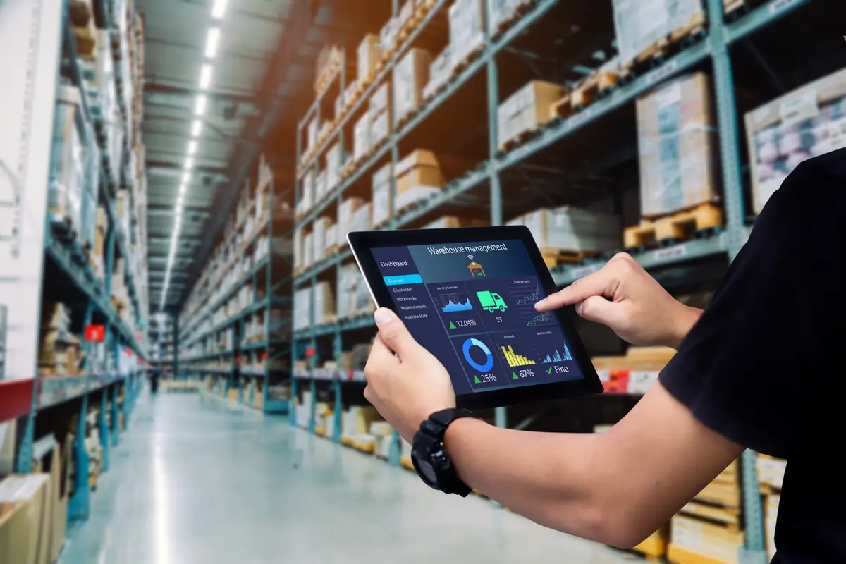 Mão de uma pessoa segurando um tablet com tela de gestão de estoque em um grande depósito