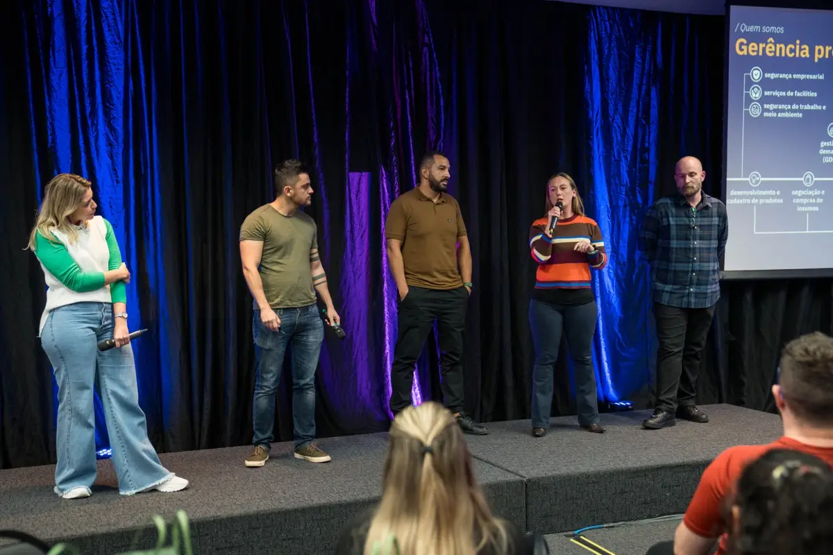 Cinco pessoas em um palco com microfones, uma mulher falando para a plateia durante apresentação.