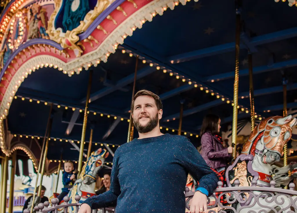 Homem com blusa azul ao lado de carrossel decorado com cavalos iluminados