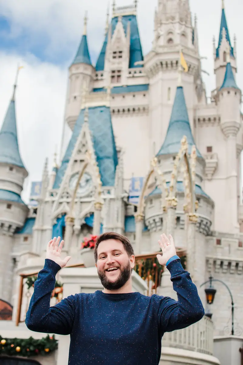 Homem sorridente com camiseta azul levantando as mãos em frente ao castelo da Disney em Orlando