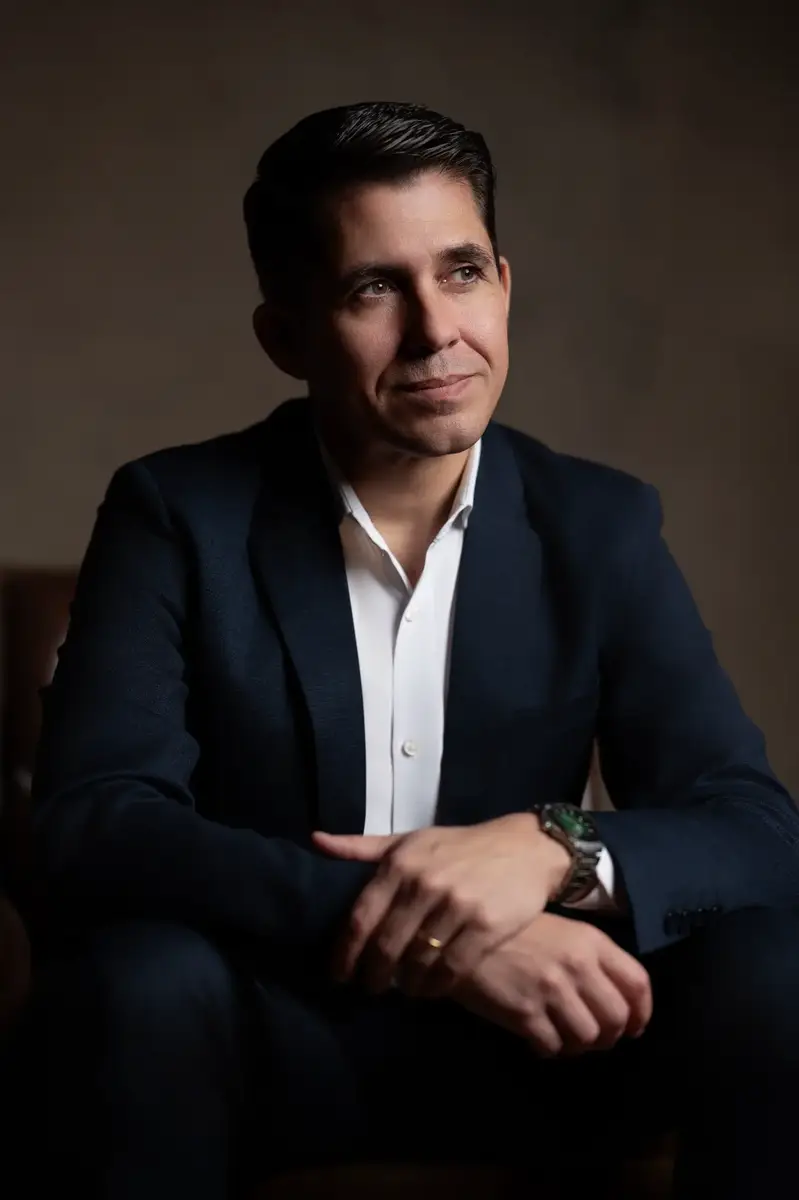 Homem vestido com terno azul escuro e camisa branca sentado, olhando levemente para o lado