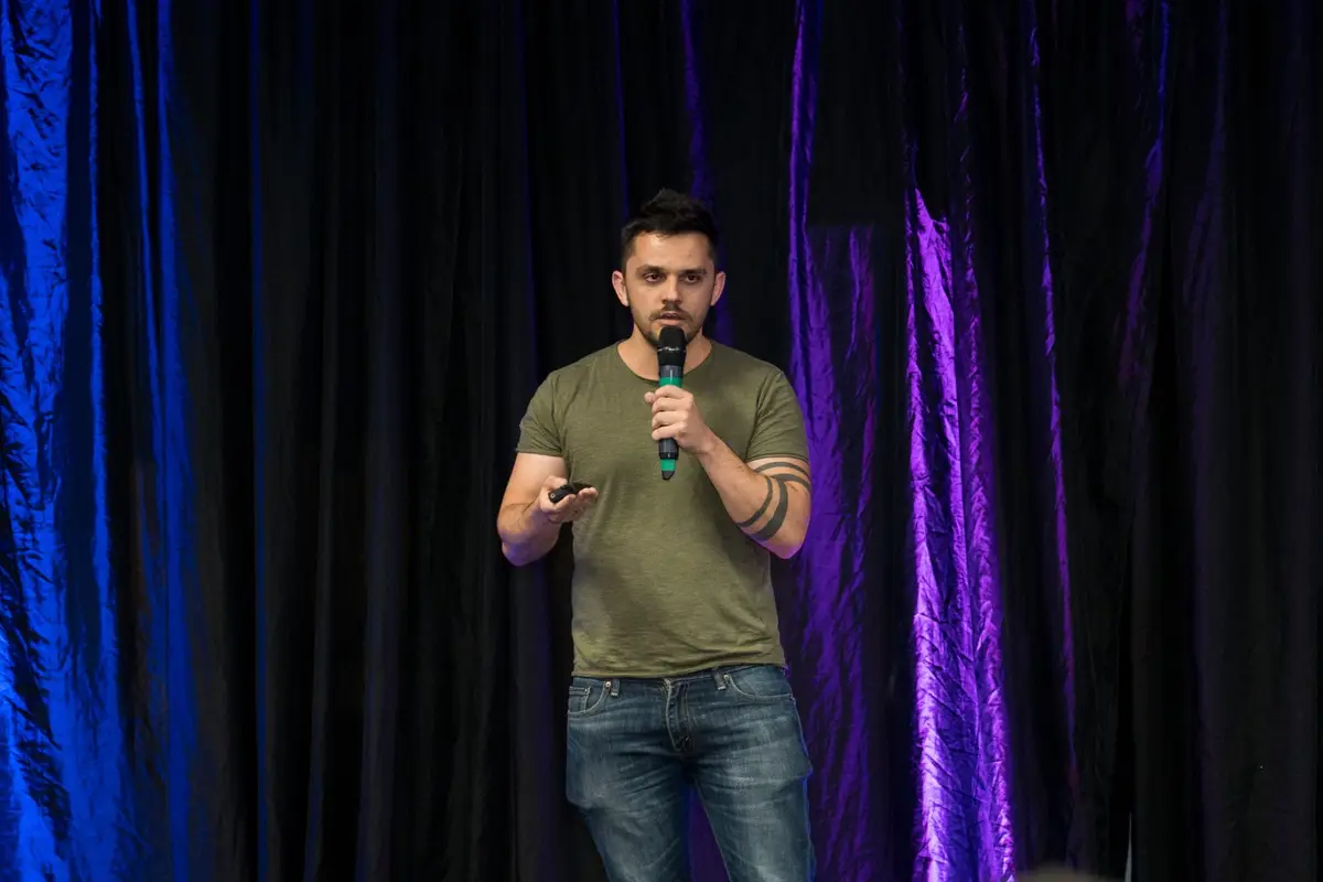 Homem jovem falando com microfone em palco com cortinas pretas e iluminação azul e roxa