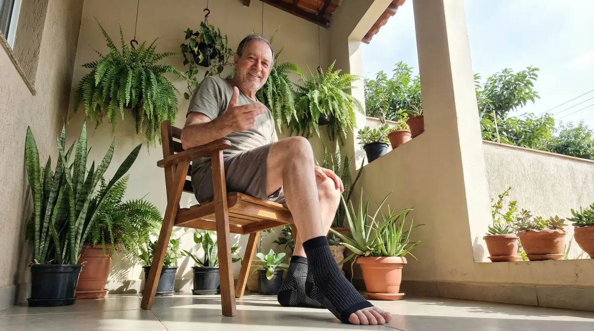 Homem adulto sentado em cadeira de madeira em varanda com várias plantas em vasos ao fundo, usando meias de compressão NeuroFit.