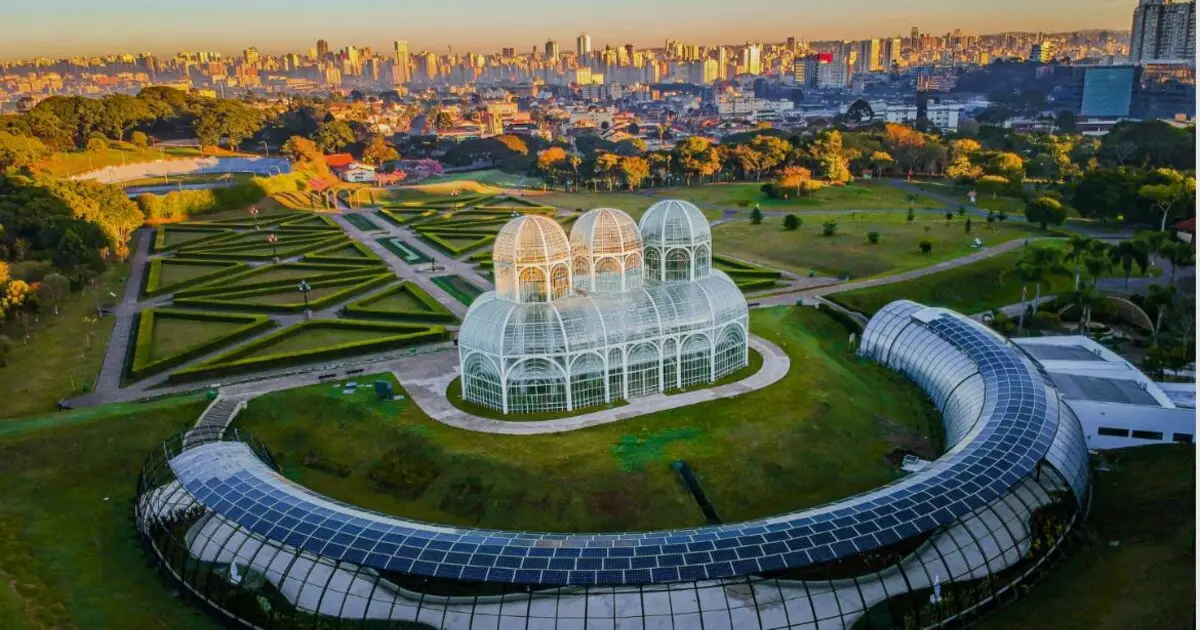 Vista aérea do Jardim Botânico de Curitiba ao entardecer com estufa de ferro e vidro e os prédios da cidade ao fundo