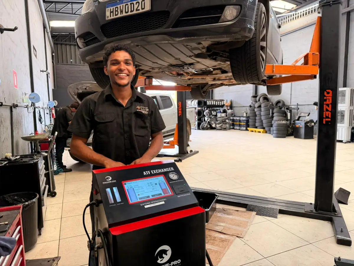 Mecânico sorridente em oficina ao lado de equipamento de troca de óleo de câmbio com carro elevado ao fundo