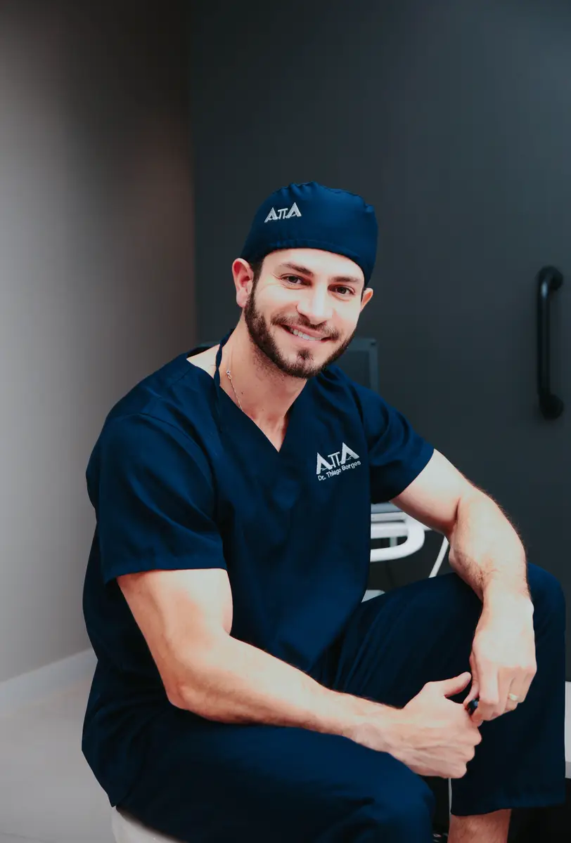 Médico com uniforme cirúrgico azul escuro sentado e sorrindo