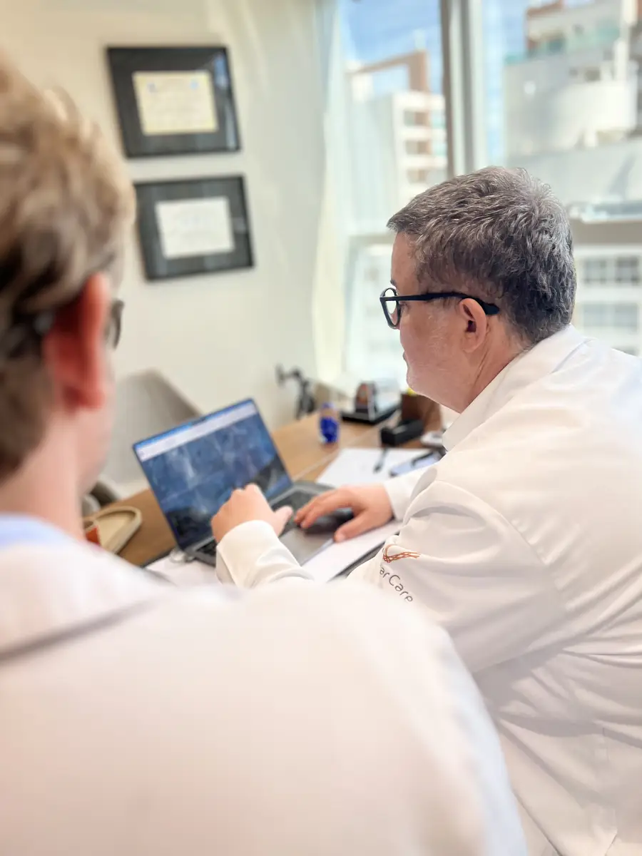 Dois médicos em jaleco consultando imagens em um laptop em consultório com janelas amplas ao fundo