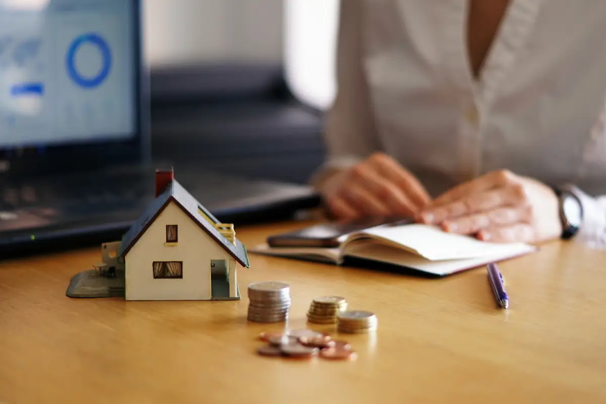 Miniatura de casa com moedas empilhadas e pessoa escrevendo em caderno ao fundo