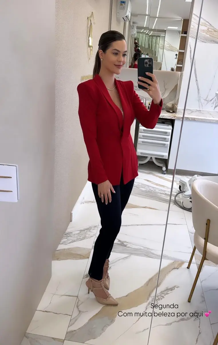 Mulher vestindo blazer vermelho e calça preta tirando selfie em espelho em clínica estética