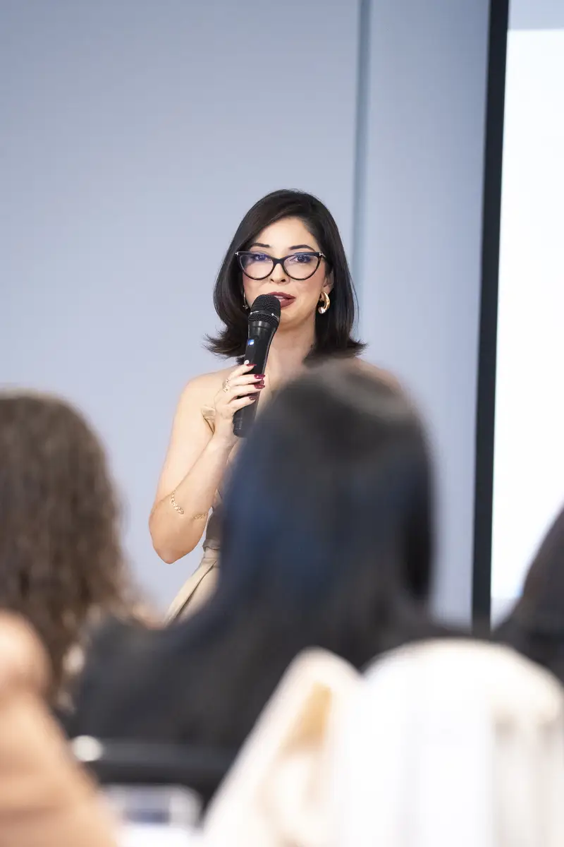 Mulher de óculos falando ao microfone para uma audiência em ambiente de apresentação