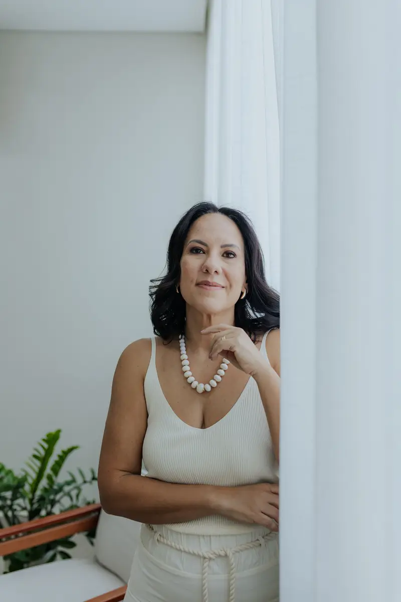 Mulher de meia-idade com cabelo preto e ondulado, usando regata branca e colar de contas brancas, apoiada em cortina branca em ambiente iluminado