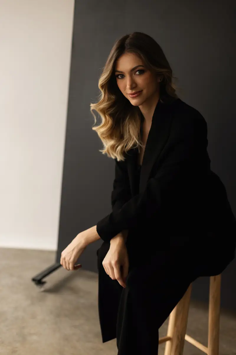 Mulher sentada em banquinho de madeira com cabelo loiro ondulado e blazer preto