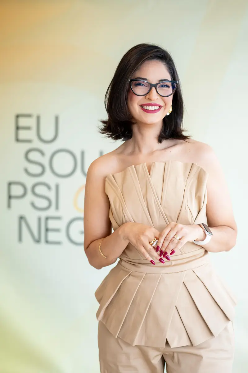 Mulher sorridente com blusa bege sem mangas e óculos de grau em fundo claro com texto desfocado