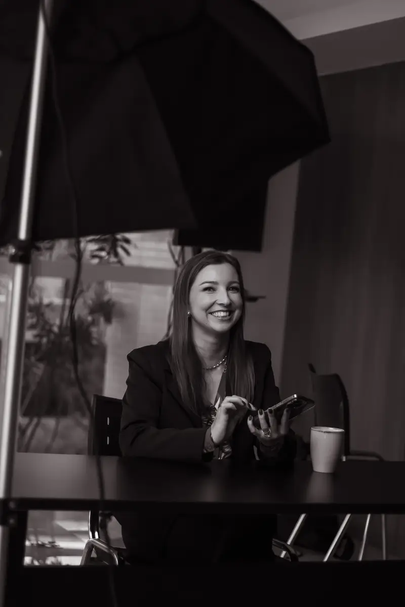 Cris Klein sorrindo sentada à mesa usando celular em ambiente de escritório