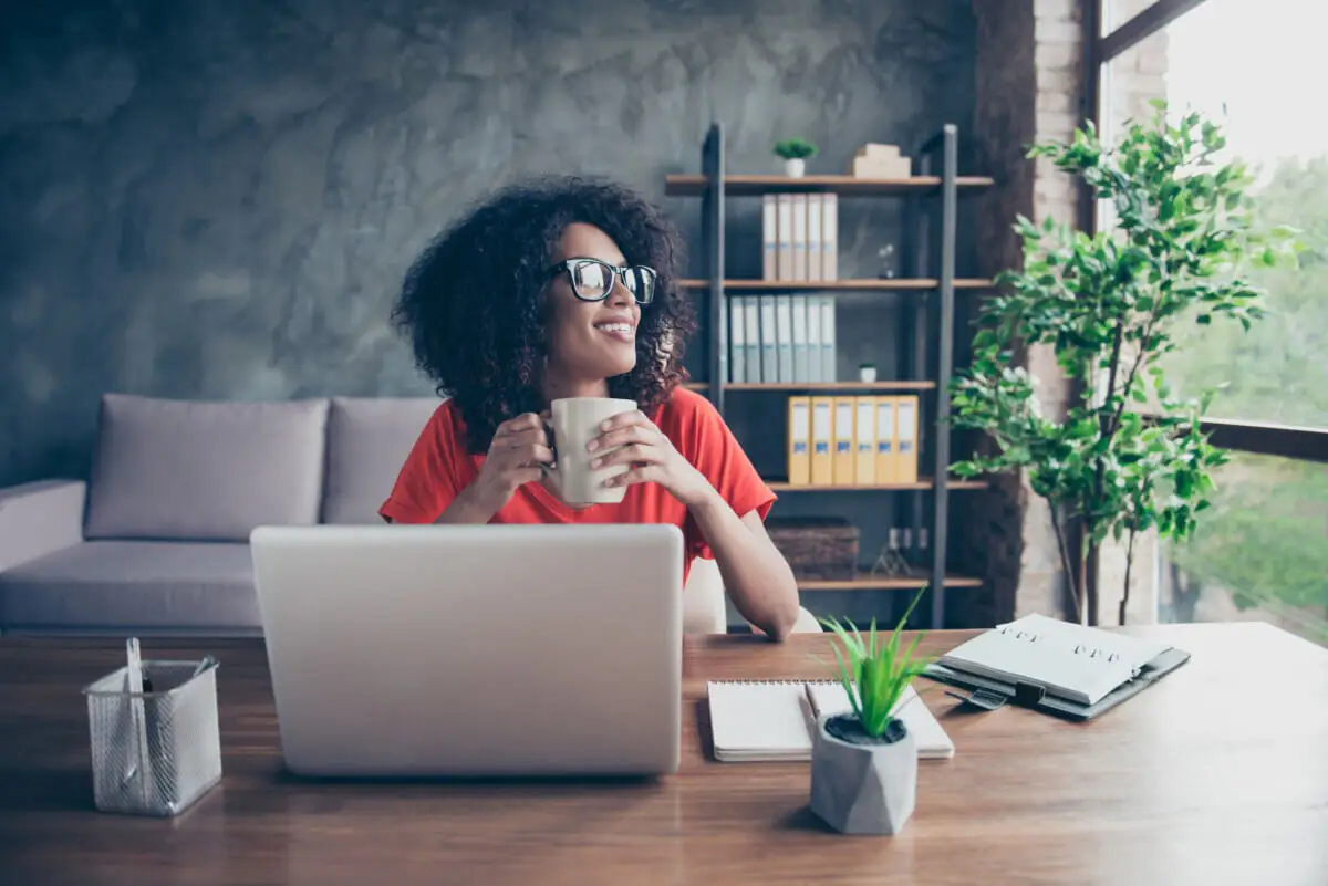 Mulher sorridente segurando uma caneca enquanto está sentada diante de um laptop em um ambiente de escritório moderno