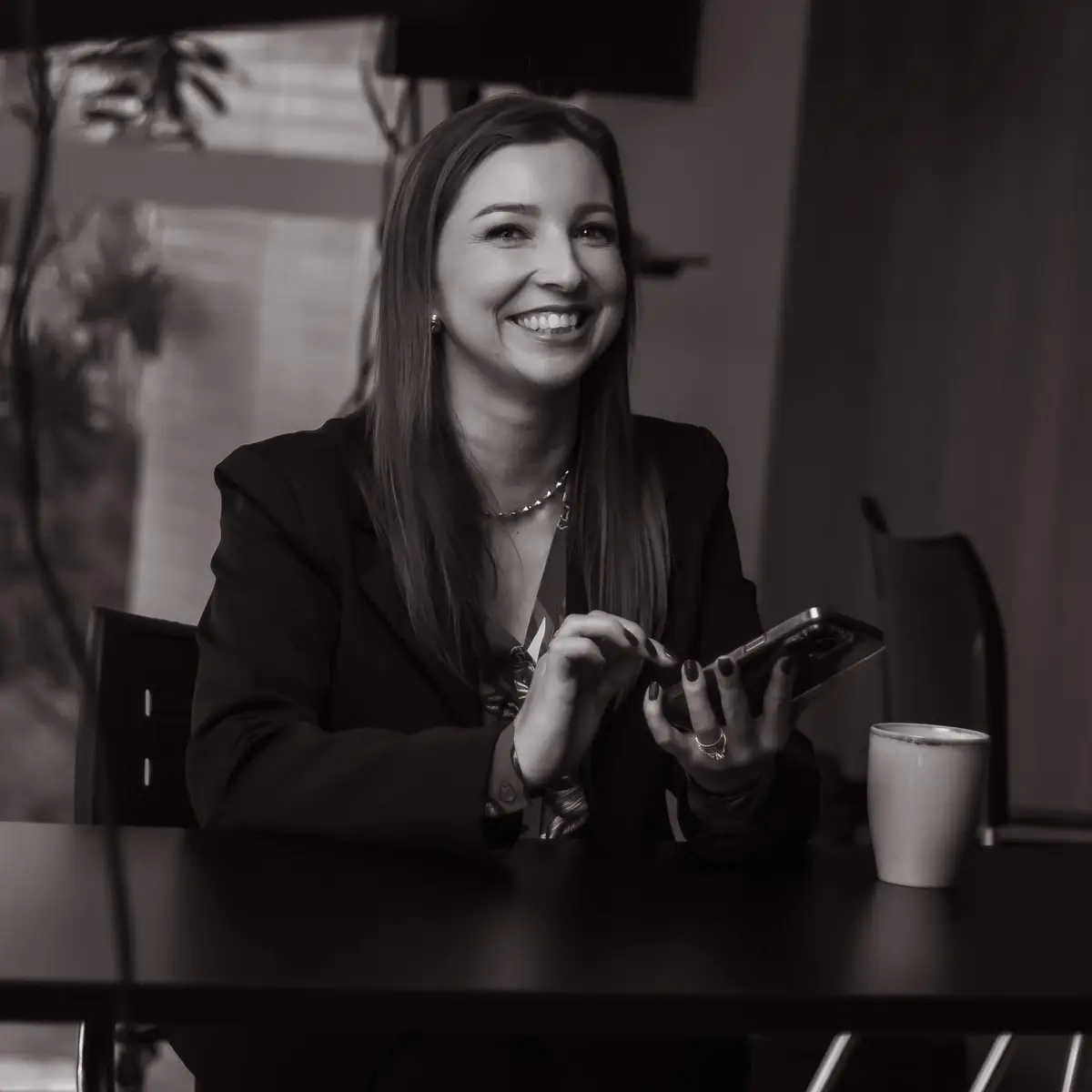 Mulher sorridente usando celular sentada à mesa com caneca ao lado