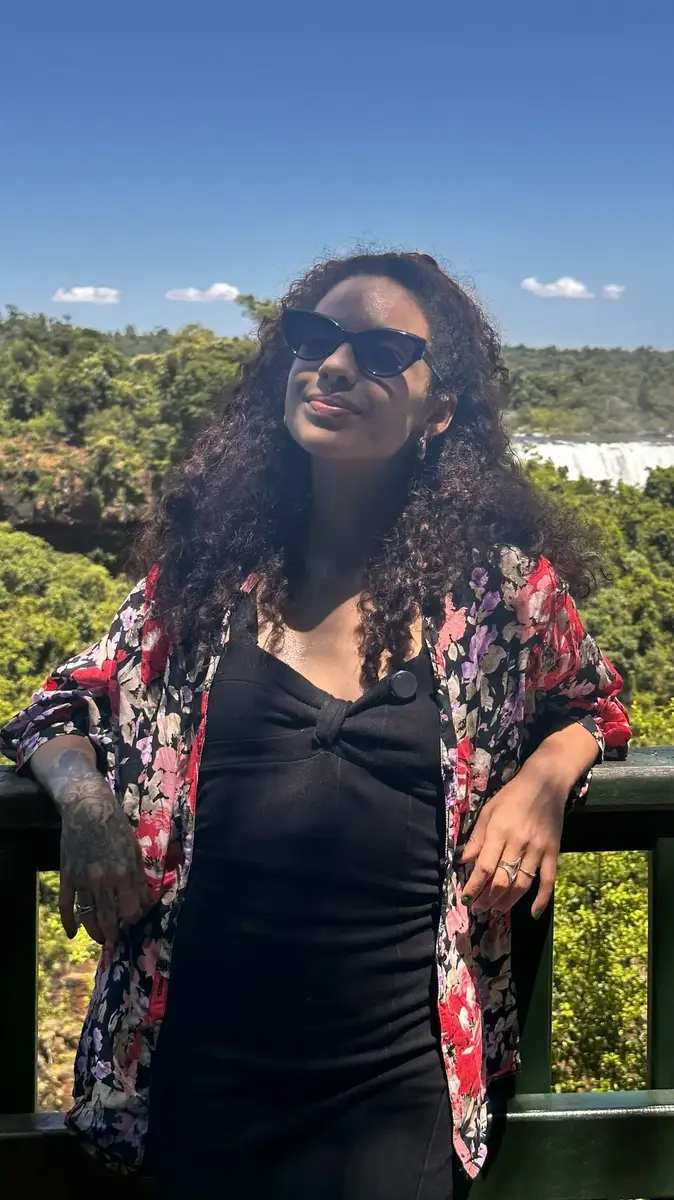 Mulher com vestido preto e camisa floral encostada em guarda-corpo com cataratas ao fundo
