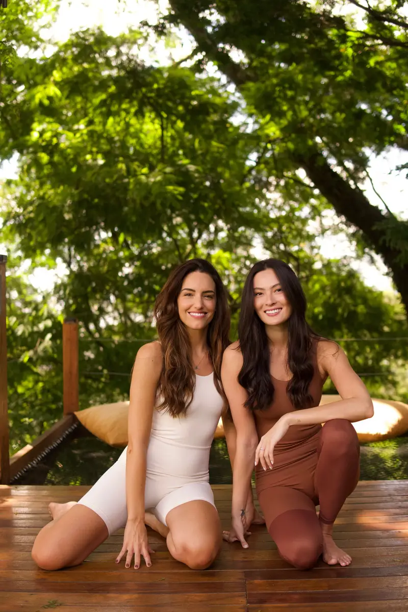 Duas mulheres sorridentes em roupas de ginástica sentadas no deque de madeira com vegetação verde ao fundo