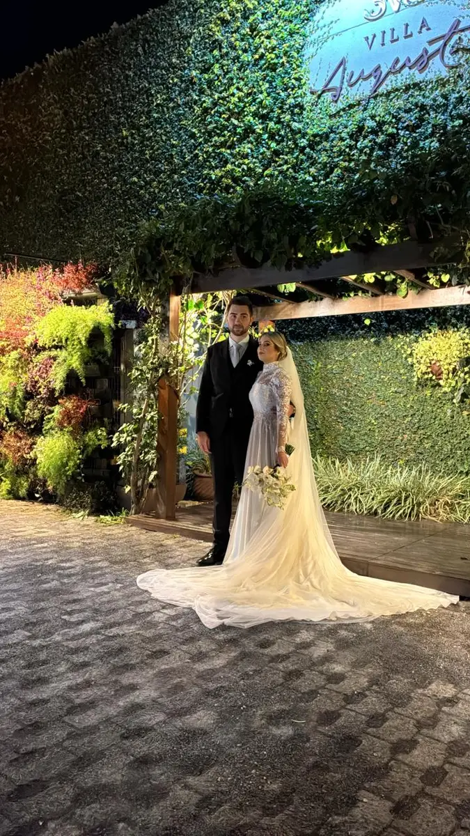 Noivos em traje formal posando em área externa com parede verde iluminada à noite