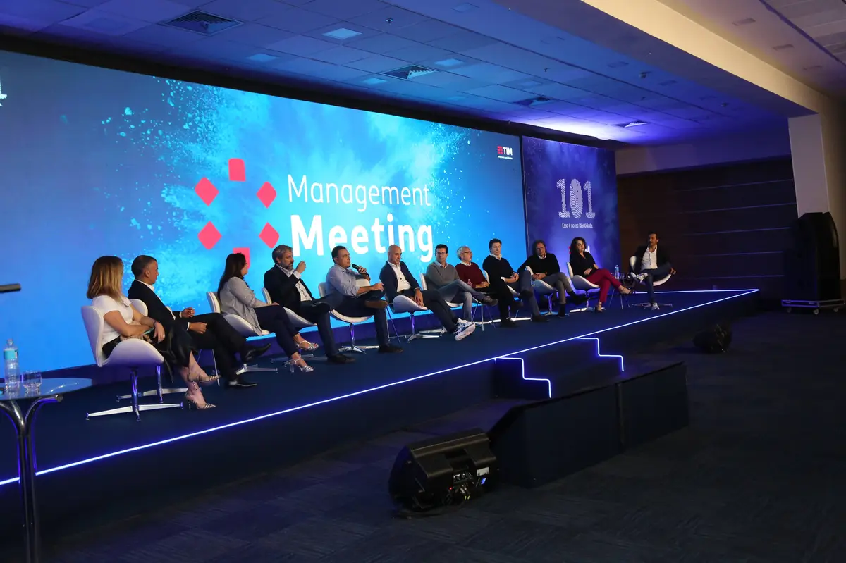 Painel de discussão com dez pessoas sentadas em cadeiras em um palco iluminado azul.