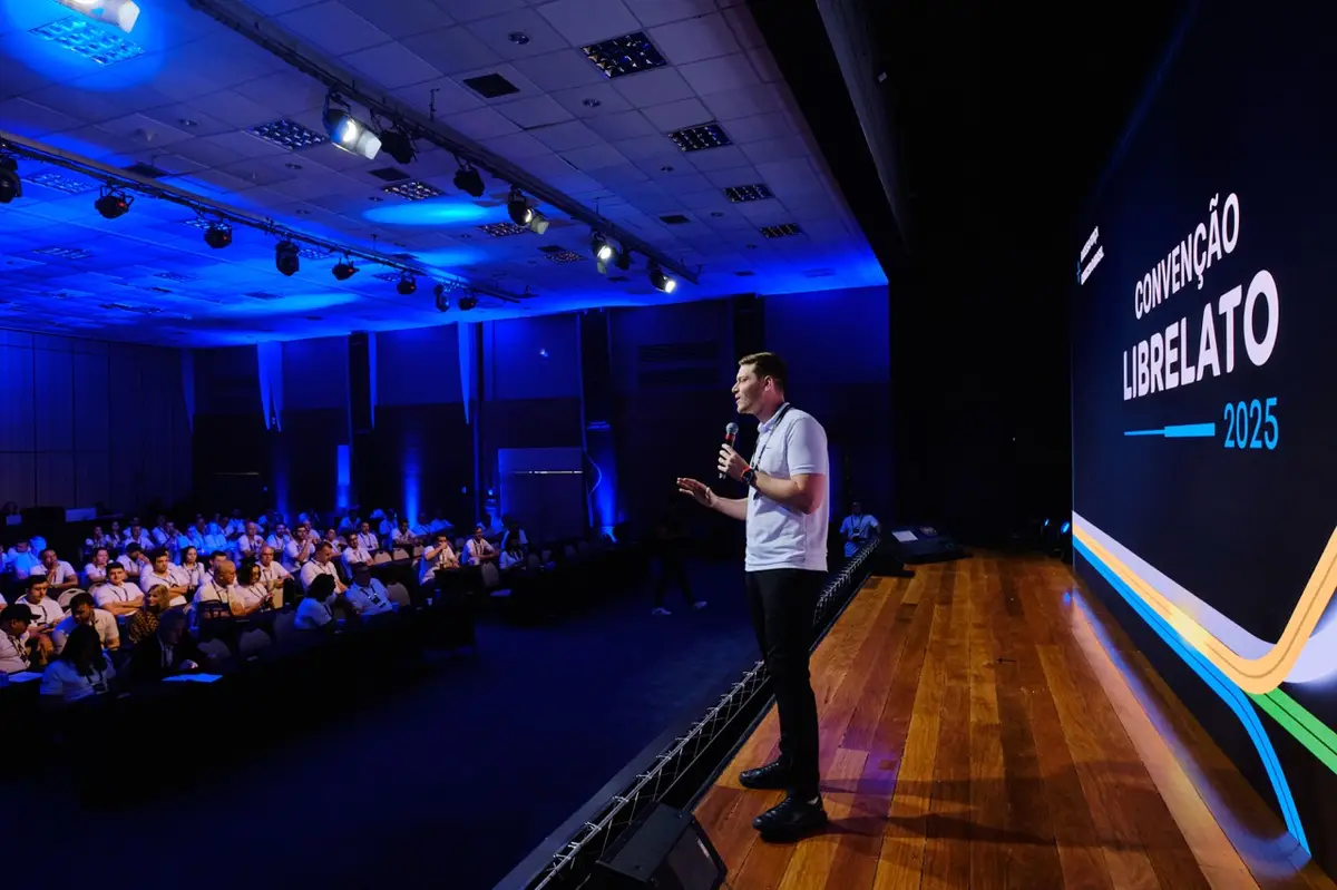 Homem falando em um palco durante convenção com público sentado em auditório azul