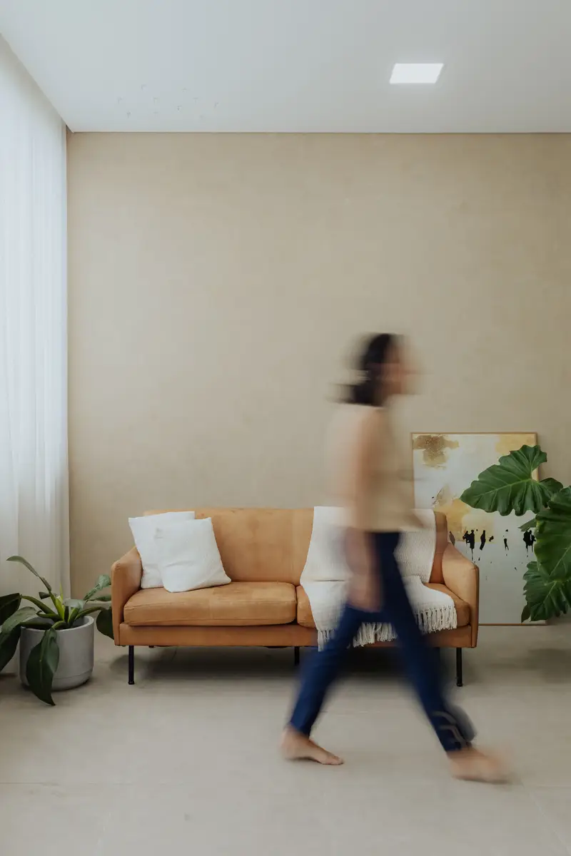 Pessoa caminhando descalça em um ambiente interno claro com sofá marrom e plantas