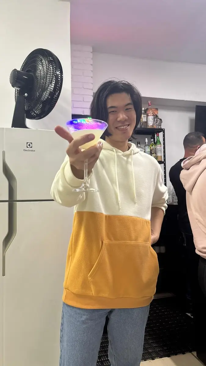 Pessoa sorrindo com moletom branco e amarelo segurando taça com drink iluminado por luzes coloridas