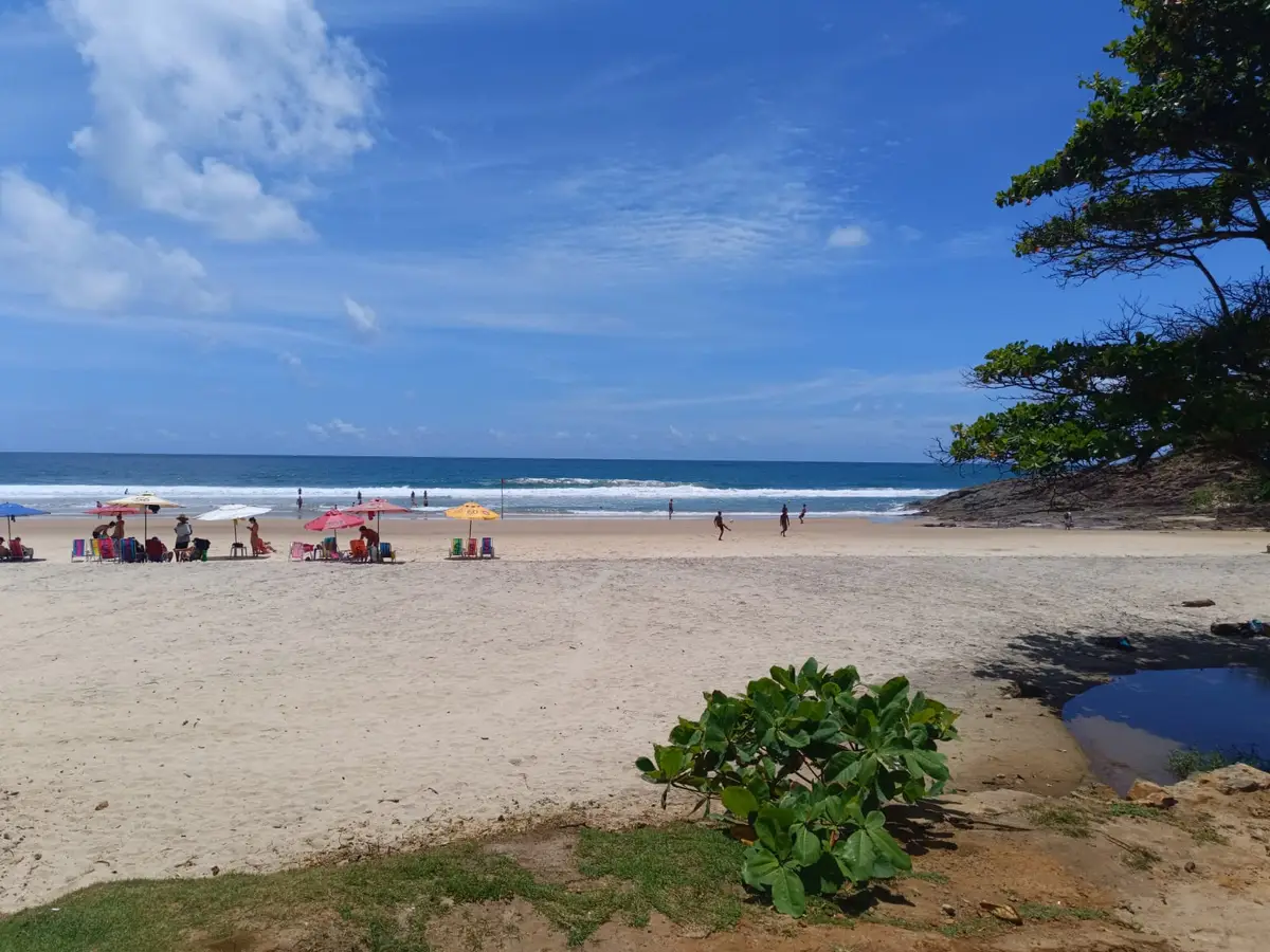 Praia de areia clara com guarda-sóis coloridos e pessoas na beira do mar azul sob céu parcialmente nublado