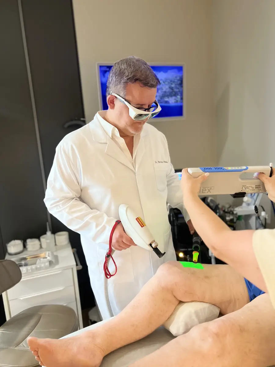 Dr Marcelo Mandelli com jaleco branco e óculos de proteção realizando procedimento a laser na perna de paciente em consultório.