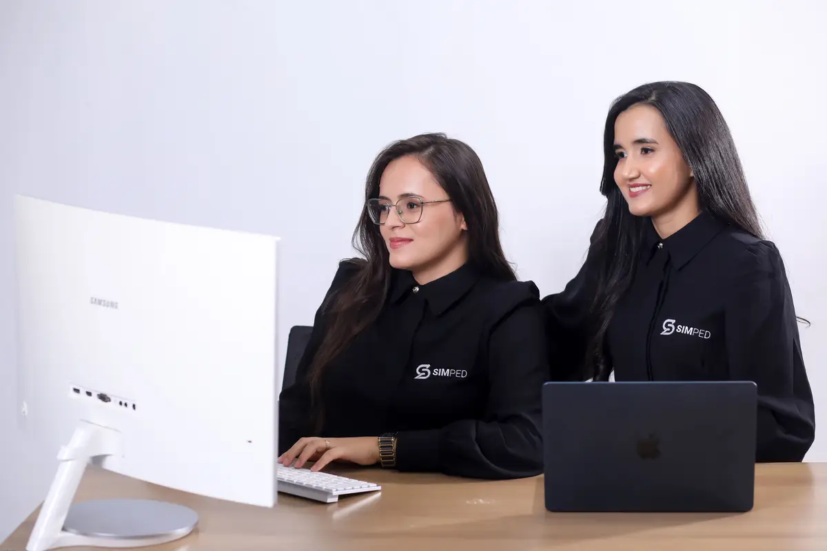 Duas mulheres usando camisas pretas com o logo da Simped trabalhando juntas em um computador e um notebook em uma mesa de madeira clara.