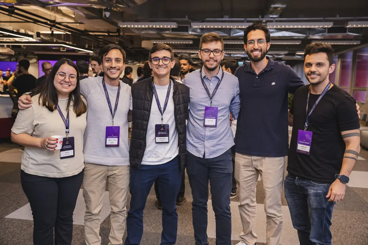 Grupo de seis profissionais sorrindo em ambiente corporativo com crachás de evento.