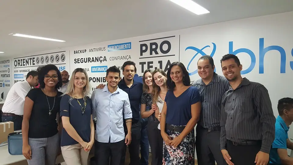 Grupo de profissionais técnicos posando para foto em ambiente de trabalho com mural de palavras relacionadas à tecnologia ao fundo
