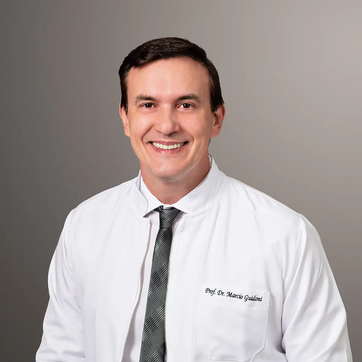 Retrato de um homem sorridente usando jaleco branco com nome bordado e gravata prata