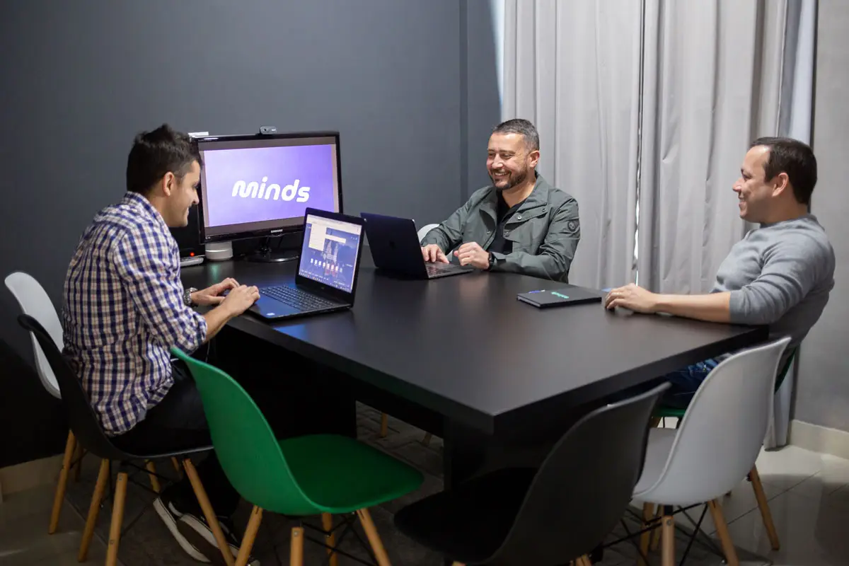 Três homens sentados em torno de uma mesa preta com laptops em uma reunião em escritório moderno