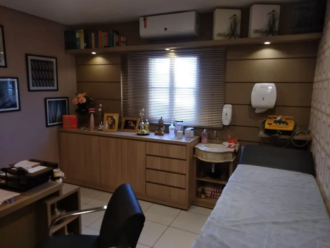 Sala de atendimento médico com maca coberta por lençol branco, cadeira preta giratória e armário de madeira sob janela com persiana