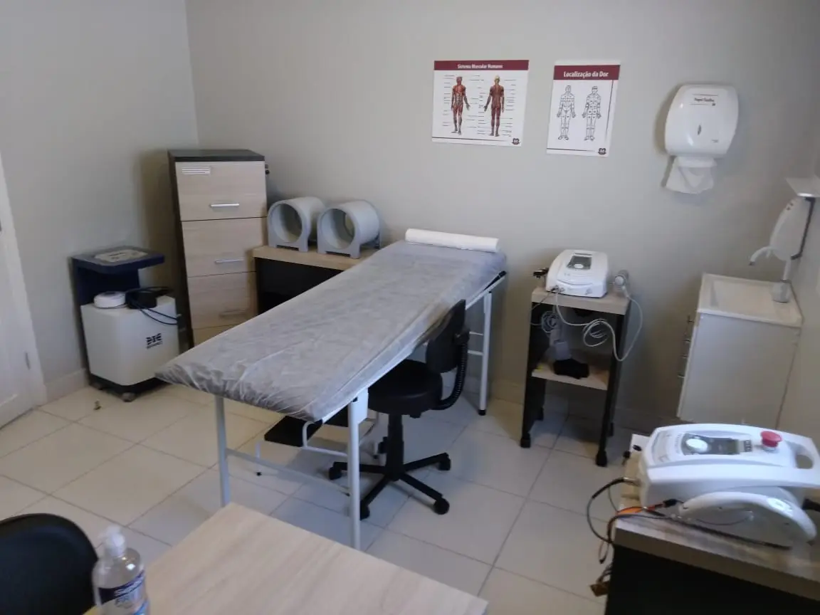 Sala de clínica médica com maca coberta por lençol, cadeira giratória, equipamentos médicos e cartazes de anatomia muscular e localização da dor na parede