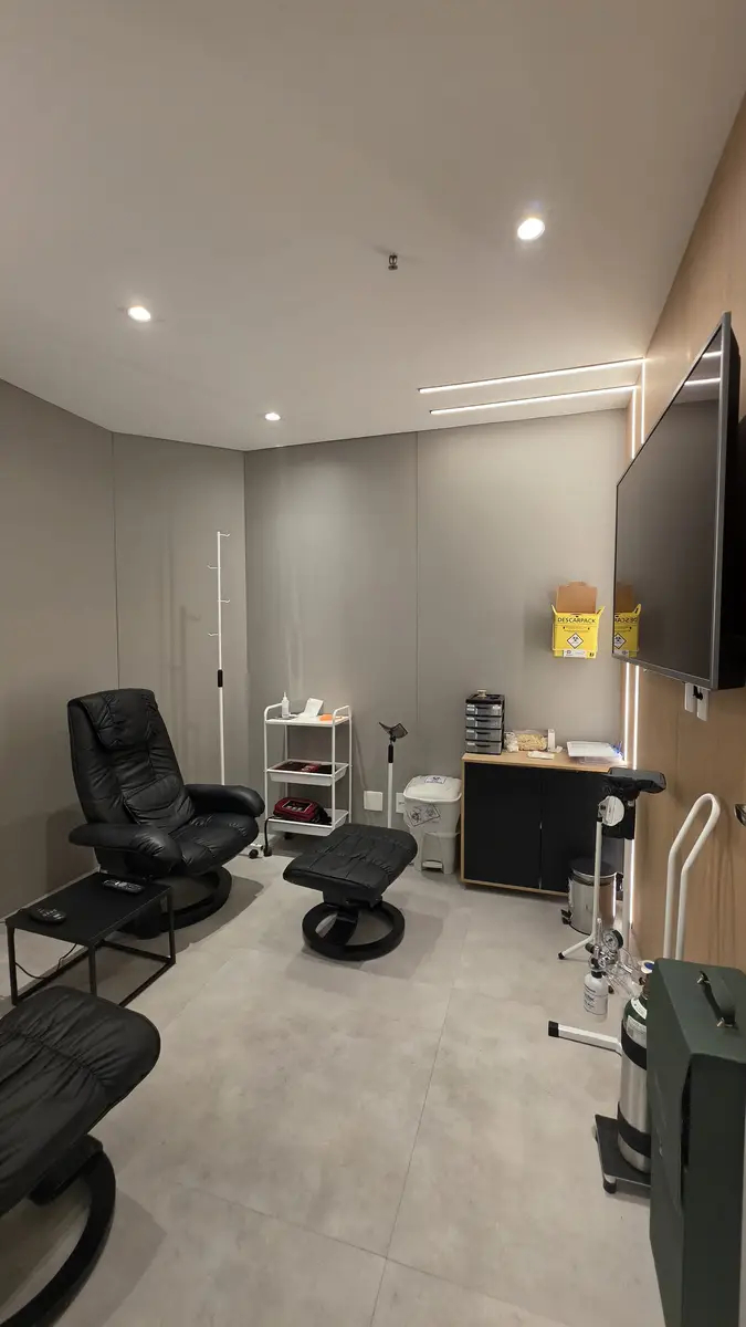 Sala de clínica com poltrona reclinável preta, estante com material médico e televisão na parede